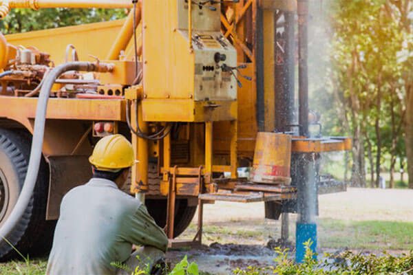 Construction worker in a yellow hard hat kneels beside a large orange drilling rig outdoors, performing maintenance or setup work.